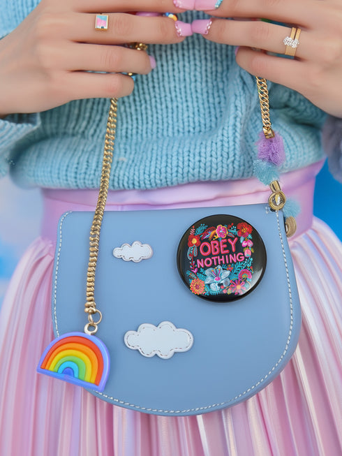 A black round pinback button featuring floral embroidery-style designs and bold red text that reads "Obey Nothing."